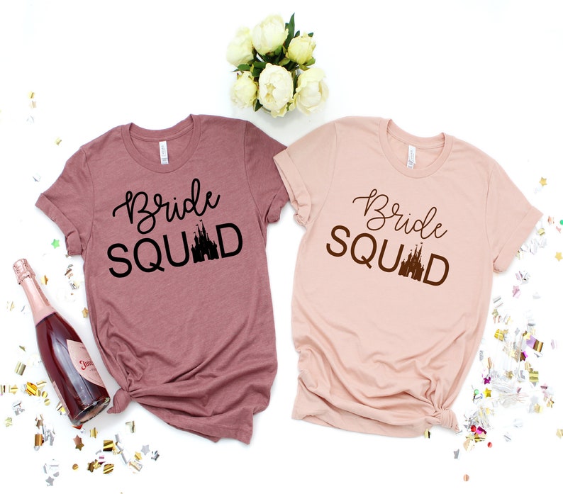 Peut inclure: Deux t-shirts avec le texte "Bride Squad" et une silhouette du ch&acirc;teau de Cendrillon. Les chemises sont dans des tons de rose et de mauve.