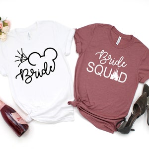 Peut inclure: Deux t-shirts blancs avec du texte noir. Le premier t-shirt dit "Bride" avec un diamant et des oreilles de Mickey Mouse. Le deuxi&egrave;me t-shirt est marron et dit "Bride Squad" avec un graphique de ch&acirc;teau blanc.