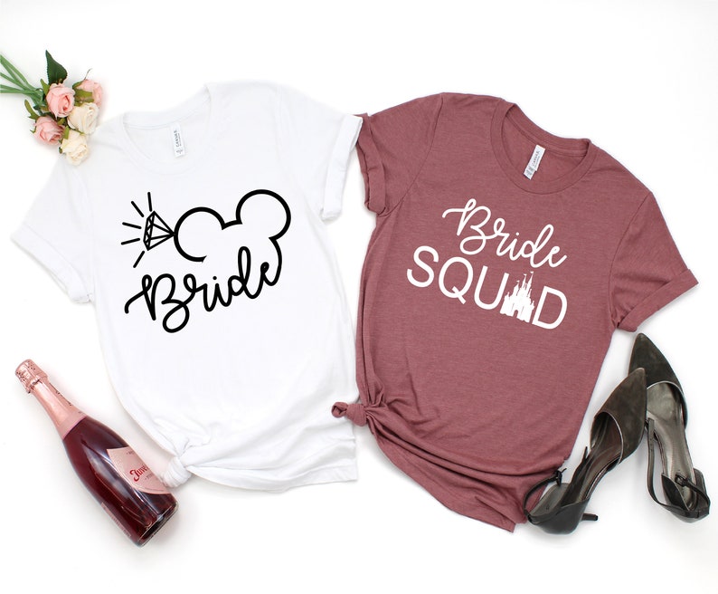 Peut inclure: Deux t-shirts blancs avec du texte noir. Le premier t-shirt dit "Bride" avec un diamant et des oreilles de Mickey Mouse. Le deuxi&egrave;me t-shirt est marron et dit "Bride Squad" avec un graphique de ch&acirc;teau blanc.