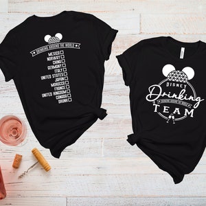 May include: Two black t-shirts with white text and graphics. One shirt says "Drinking Around the World" with a list of countries and checkboxes. The other shirt says "Disney Drinking Team" with a Mickey Mouse ear silhouette and a Spaceship Earth graphic.