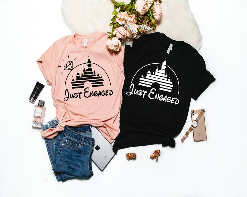 May include: Two t-shirts with a black and white graphic of Cinderella's castle with the text "Just Engaged" on each shirt. One shirt is pink and the other is black.