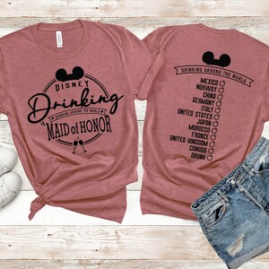 Może przedstawiać: R&oacute;żowy T-shirt z czarnym nadrukiem, na kt&oacute;rym widnieje napis "Disney Drinking" z głową Myszki Miki i tekst "Drinking Around the World" w k&oacute;łku. Pod k&oacute;łkiem znajduje się napis "Maid of Honor". Na plecach T-shirtu znajduje się czarny nadruk z napisem "Drinking Around the World" z głową Myszki Miki i listą kraj&oacute;w.