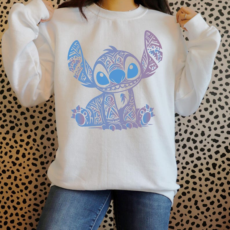 K&ouml;nnte beinhalten: Ein wei&szlig;er Pullover mit einem blauen und violetten Stitch-Charakter-Motiv. Der Charakter sitzt mit gekreuzten Armen und hat ein blaues und violettes Muster.