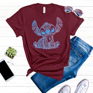 K&ouml;nnte beinhalten: Ein maroonfarbenes T-Shirt mit einem blauen und wei&szlig;en tribalen Stitch-Muster. Das Shirt ist gefaltet und auf einer wei&szlig;en Oberfl&auml;che mit Jeans, Sonnenbrille und Sneakern pr&auml;sentiert.