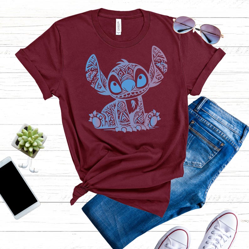 K&ouml;nnte beinhalten: Ein maroonfarbenes T-Shirt mit einem blauen und wei&szlig;en tribalen Stitch-Muster. Das Shirt ist gefaltet und auf einer wei&szlig;en Oberfl&auml;che mit Jeans, Sonnenbrille und Sneakern pr&auml;sentiert.