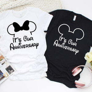 May include: Two t-shirts, one white and one black, with the text "It's Our Anniversary". The white shirt features a black bow and mouse ears, while the black shirt has white mouse ears. The shirts are casual wear.