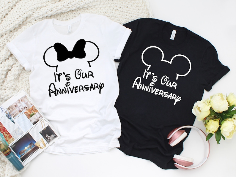 May include: Two t-shirts, one white and one black, with the text "It's Our Anniversary". The white shirt features a black bow and mouse ears, while the black shirt has white mouse ears. The shirts are casual wear.