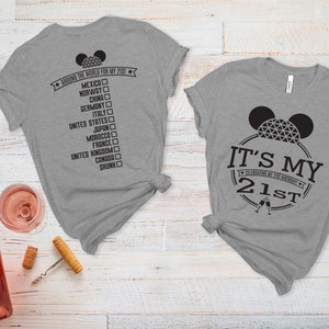 May include: Two gray t-shirts with black text and graphics. The first shirt says "Around the world for my 21st" with a list of countries and checkboxes. The second shirt says "It's my 21st" with a Mickey Mouse head and a champagne glass graphic.