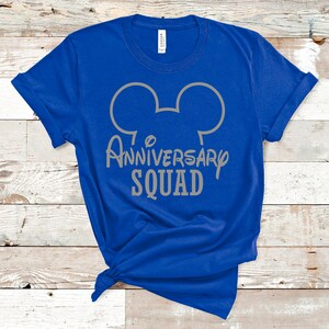 May include: A bright blue t-shirt with a grey Mickey Mouse head outline and the words "Anniversary Squad". The shirt is knotted at the bottom. The background is a light wood-grain surface.