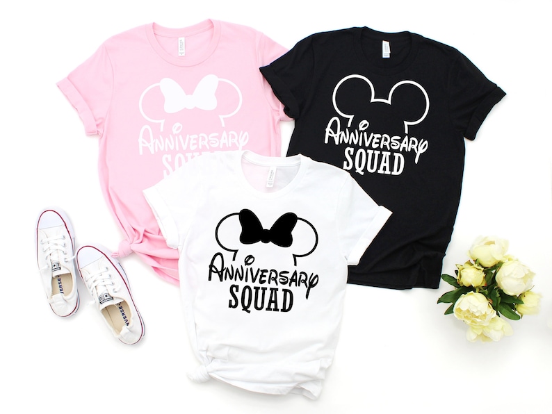 May include: Three t-shirts in pink, white, and black, each with a Mickey Mouse-style design, a bow, and the text "Anniversary Squad." White trainers are near the pink and white shirts. A bouquet of white flowers is next to the black shirt.