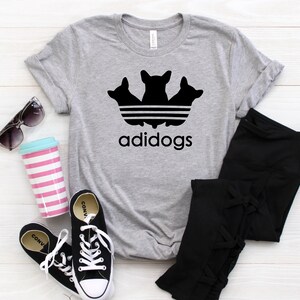 Puede incluir: Una camiseta gris con un gráfico negro de tres perros en un logotipo de Adidas estilizado. El texto "adidogs" está impreso debajo del logotipo.