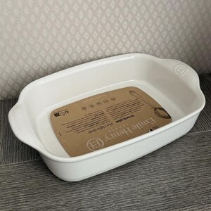 May include: A rectangular white ceramic baking dish with rounded corners and integrated side handles. A brown paper product label is placed inside the dish, featuring the text Emile Henry, le bon plat, and the right dish, along with icons.