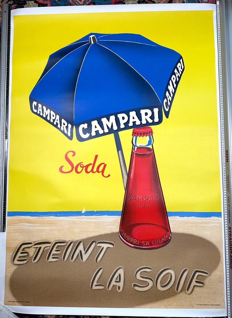 Pode incluir: Um an&uacute;ncio vintage de Campari Soda. A imagem mostra um guarda-sol de praia azul e branco listrado com a palavra "Campari" impressa nele. Uma garrafa de Campari Soda est&aacute; sentada em uma praia de areia com as palavras "Eteint La Soif" escritas na areia.