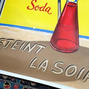 Pode incluir: Um an&uacute;ncio vintage de Campari Soda. A imagem apresenta uma garrafa de Campari Soda numa praia com as palavras "Eteint La Soif" escritas na areia. O fundo &eacute; amarelo brilhante com um oceano azul e veleiros brancos.