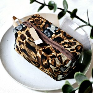 K&ouml;nnte beinhalten: Eine braune und beige gepunktete Rei&szlig;verschlusstasche mit einem braunen Rei&szlig;verschluss und einem beigefarbenen Stoffanh&auml;nger. Die Tasche liegt auf einem wei&szlig;en Teller.