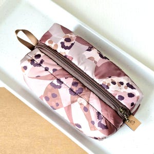 Könnte beinhalten: Eine rosa und braune Reißverschlusstasche mit Leopardenmuster. Die Tasche hat einen braunen Reißverschluss und einen braunen Riemen.