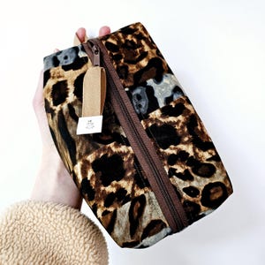 K&ouml;nnte beinhalten: Eine braune und schwarze Leopardenmuster-Rei&szlig;verschlusstasche mit einem beigefarbenen Rei&szlig;verschluss-Zipper. Die Tasche ist aus Stoff und hat ein Patchwork-Design.
