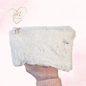 Könnte beinhalten: Eine weiße, flauschige, mit einem Reißverschluss versehene Tasche mit einem kleinen weißen Etikett, auf dem "Design" steht.
