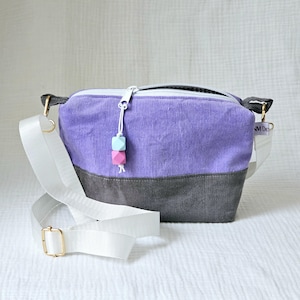 Könnte beinhalten: Eine kleine Umhängetasche aus violettem und grauem Cord mit einem weißen Trageriemen und einem Reißverschluss. Die Tasche hat einen pinkfarbenen und blauen Holzperlen-Anhänger, der am Reißverschluss befestigt ist.
