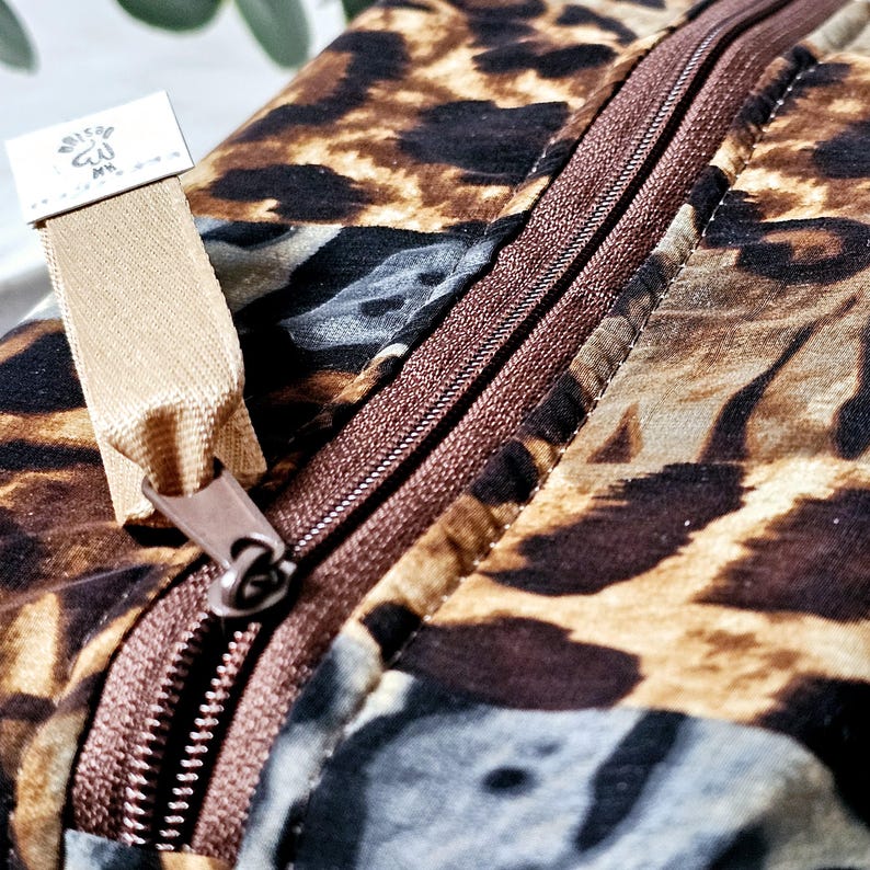K&ouml;nnte beinhalten: Eine braune und beige gepunktete Rei&szlig;verschlusstasche mit einem braunen Rei&szlig;verschluss und einem beigefarbenen Stoffzugband. Die Tasche hat ein kleines wei&szlig;es Etikett mit dem Text "Blissful" und einem Symbol.