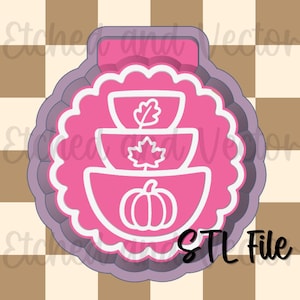May include: A pink cookie cutter with a scalloped edge featuring a stacked design of three bowls. The top bowl contains a leaf, the middle bowl contains a maple leaf, and the bottom bowl contains a pumpkin. The text STL File is visible.