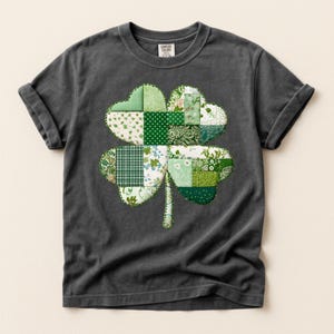 Può includere: T-shirt grigio scuro con un design a trifoglio patchwork. Il trifoglio è composto da vari tessuti verdi con motivi floreali, a pois e a quadri. La maglietta ha maniche arrotolate e girocollo.
