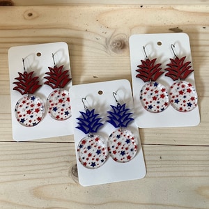 May include: Three pairs of dangle earrings featuring a pineapple design with red, white, and blue star accents. The pineapple is made of wood and the star accents are on a clear acrylic disc.
