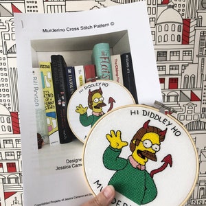 Op de afbeelding: Een borduurring met kruissteek met een witte achtergrond en een geel cartoonfiguur met een groen shirt en rode duivelshoorns. Het figuur zwaait en de tekst "Hi Diddley Ho Murderino" staat rondom het figuur.