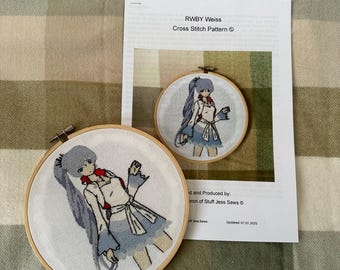 May include: Two cross-stitch embroidery hoops featuring a character with long, light blue hair, a white outfit, and a blue skirt. The text "RWBY Weiss Cross Stitch Pattern" is visible on a printed sheet.