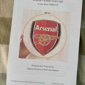 May include: A cross-stitch pattern of the Arsenal Football Club logo, featuring a red and white shield with the word "Arsenal" and a cannon. The pattern is held in a wooden embroidery hoop. The text "Arsenal Football Club Logo Cross Stitch Pattern" is printed above the design.