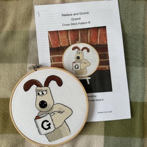 Könnte beinhalten: Ein fertiger Kreuzstich-Stickrahmen mit Gromit aus Wallace und Gromit, der eine Tasse mit dem Buchstaben "G" darauf hält. Der Rahmen ist aus Holz und hat einen weißen Stoffhintergrund. Das Bild stammt aus einem Kreuzstichmuster mit dem Titel "Wallace und Gromit: Gromit" von Jess Sews.