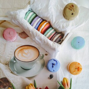 Può includere: Immagine di natura morta con macarons colorati, una tazza di caffè con latte art e tulipani vibranti. I macarons sono in vari colori, tra cui rosa, blu e arancione. La tazza di caffè è bianca e i tulipani sono rossi e gialli.