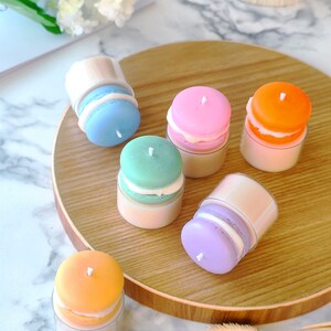 Può includere: Una collezione di candele colorate a forma di macaron nei toni del blu, rosa, verde, arancione e viola. Ogni candela ha uno stoppino bianco e si trova in un contenitore trasparente, disposte su un vassoio di legno. Decorativo e unico.