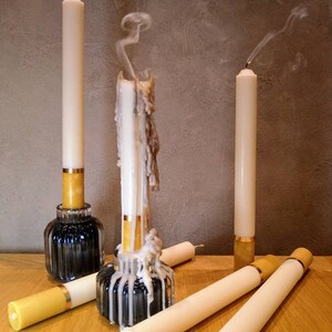 May include: A collection of ivory-colored taper candles, some extinguished with visible smoke trails. The candles are in various stages of use, with wax drippings on some. They are set in decorative holders with gold accents, on a wooden surface.