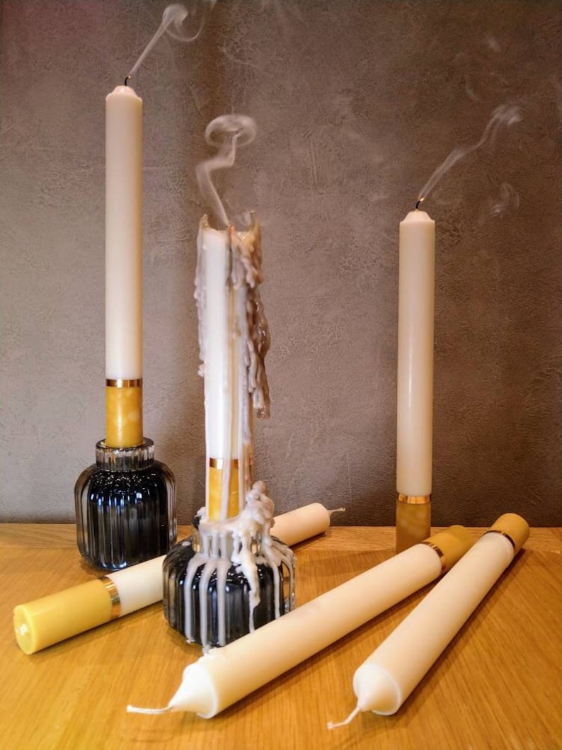 May include: A collection of ivory-colored taper candles, some extinguished with visible smoke trails. The candles are in various stages of use, with wax drippings on some. They are set in decorative holders with gold accents, on a wooden surface.