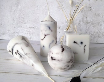 Pode incluir: Uma coleção de velas brancas com efeito mármore e veios pretos, em várias formas: um cone, uma esfera, um quadrado e um cilindro alto. As velas são dispostas com detalhes florais secos. O fundo é uma superfície branca texturizada.