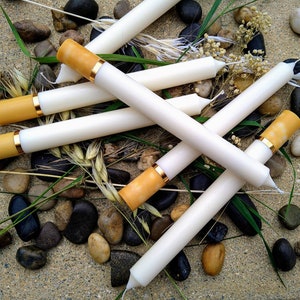 May include: A collection of white candles designed to resemble cigarettes, with tan tips and gold bands. The candles are arranged on a bed of small stones, grass, and dried flowers, creating a unique decorative display.