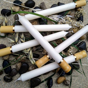 May include: White taper candles with tan tips and gold bands are arranged on a bed of pebbles, grass, and dried flowers. The candles are cylindrical and appear to be unscented. The overall aesthetic is rustic and natural.
