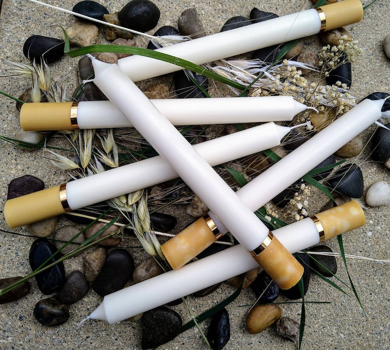 May include: White taper candles with tan tips and gold bands are arranged on a bed of pebbles, grass, and dried flowers. The candles are cylindrical and appear to be unscented. The overall aesthetic is rustic and natural.