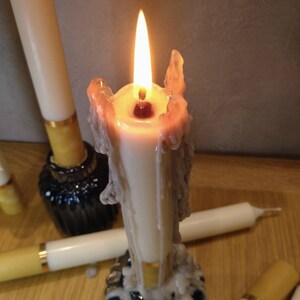 May include: A lit white candle with melted wax, surrounded by other unlit candles. The candle is in a black holder. The flame is bright yellow and orange. The candles are on a wooden surface.