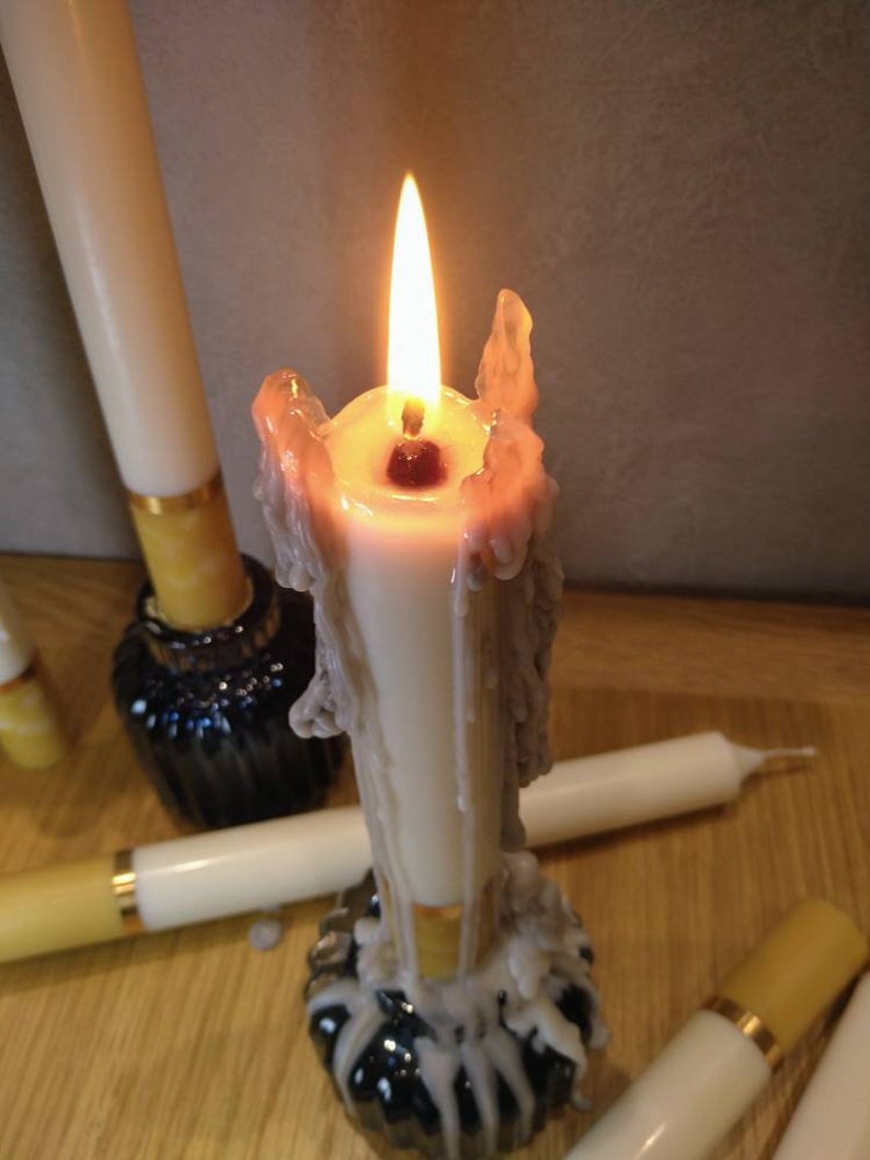 May include: A lit white candle with melted wax, surrounded by other unlit candles. The candle is in a black holder. The flame is bright yellow and orange. The candles are on a wooden surface.