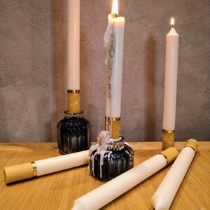May include: A collection of ivory taper candles, some lit, with gold and black accents. The candles are in black glass holders and resting on a wooden surface. The lit candles have melted wax dripping down.