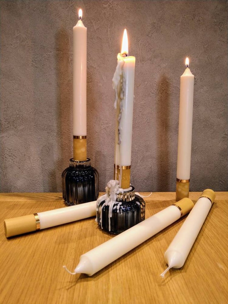 May include: A collection of ivory taper candles, some lit, with gold and black accents. The candles are in black glass holders and resting on a wooden surface. The lit candles have melted wax dripping down.