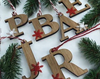 May include: Wooden Christmas ornaments spelling out the word "PEACE JR" with red and white string. The letters are brown with red and white snowflake accents. Green pine branches are in the background.