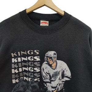 K&ouml;nnte beinhalten: Ein schwarzes Vintage-Sweatshirt mit einer Hockey-Spieler-Grafik und dem Wort "KINGS" in einem gestapelten Design. Das Sweatshirt hat einen Rundhalsausschnitt und h&auml;ngt an einem Holzkleiderb&uuml;gel.
