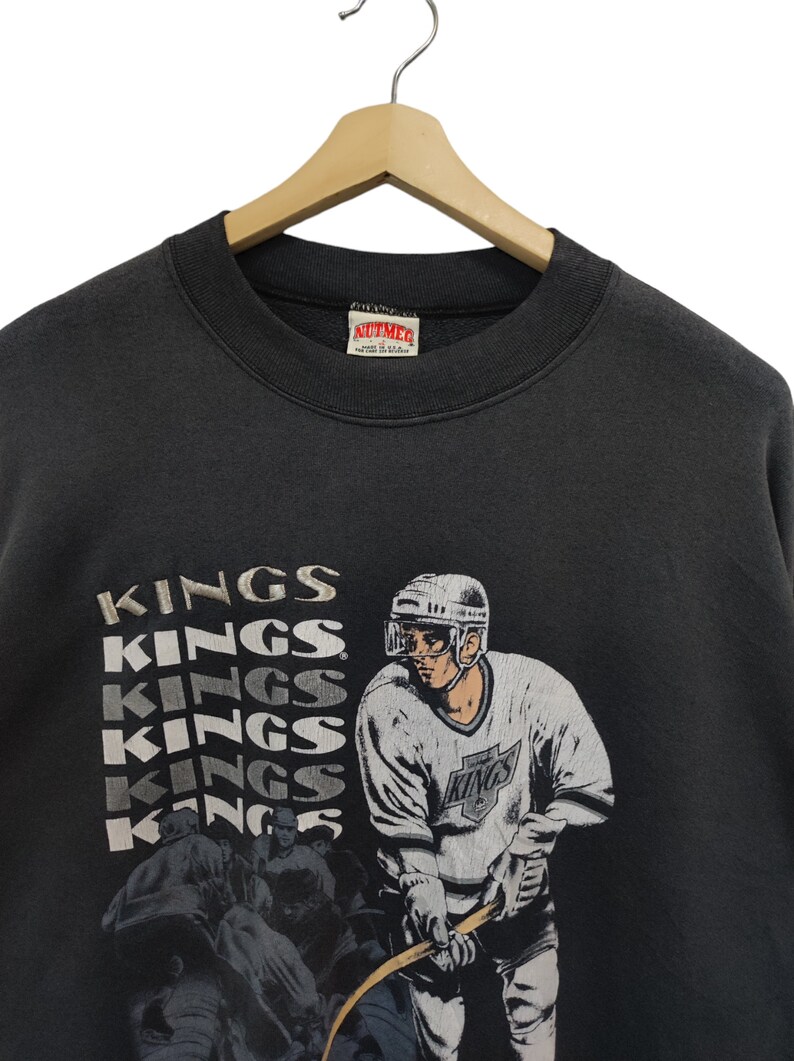 K&ouml;nnte beinhalten: Ein schwarzes Vintage-Sweatshirt mit einer Hockey-Spieler-Grafik und dem Wort "KINGS" in einem gestapelten Design. Das Sweatshirt hat einen Rundhalsausschnitt und h&auml;ngt an einem Holzkleiderb&uuml;gel.