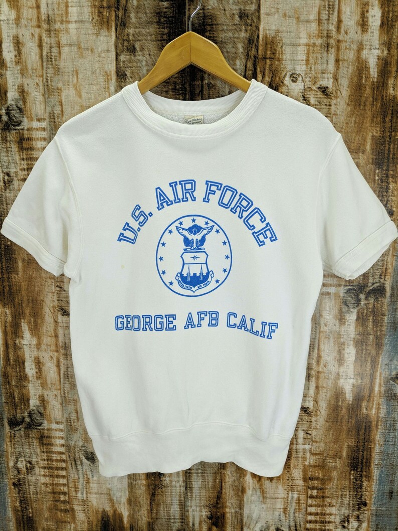 May include: White short-sleeved sweatshirt with blue text and graphics. The text reads "U.S. AIR FORCE" above a circular emblem. Below the emblem, the text "GEORGE AFB CALIF" is printed in blue.