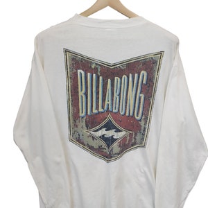 May include: A white long-sleeved shirt with a distressed graphic on the back. The graphic features the word "BILLABONG" in blue and yellow, with a diamond-shaped logo below. The shirt is hanging on a wooden hanger.