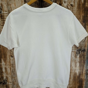 May include: A plain white short-sleeved sweatshirt hangs on a wooden hanger. The crew neck and short sleeves are visible. The sweatshirt is made of a soft, thick material. The background is a wooden surface.