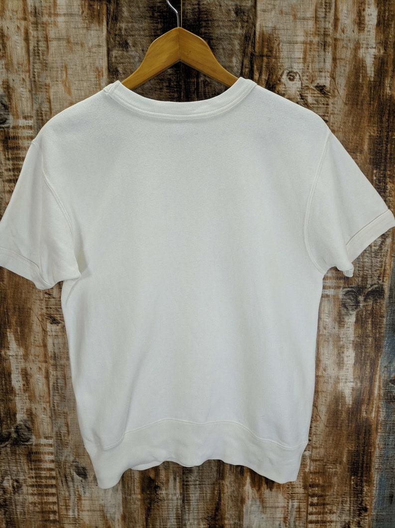 May include: A plain white short-sleeved sweatshirt hangs on a wooden hanger. The crew neck and short sleeves are visible. The sweatshirt is made of a soft, thick material. The background is a wooden surface.